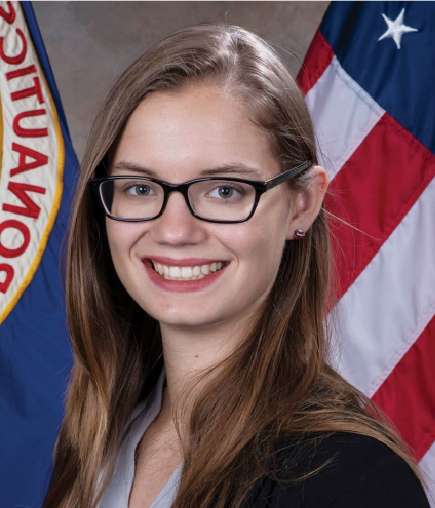 A young woman with long brown hair and glasses smiles at the camera. She is wearing a black blazer and stands in front of a U.S. flag and another banner with partially visible text.