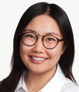 A woman with straight black hair, wearing round tortoiseshell glasses and a white collared shirt, smiles at the camera against a plain white background.