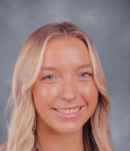 A young woman with long, wavy blonde hair smiles at the camera in front of a gray, blurred background.