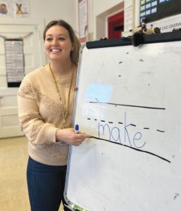 A smiling woman in a classroom points to the word make written on a whiteboard, with blank spaces and dashes before it, indicating a word or spelling activity.