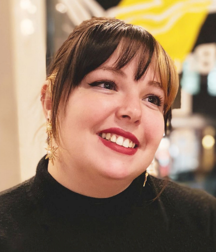 A smiling woman with dark hair and bangs, wearing a black turtleneck and gold star-shaped earrings, is photographed indoors with a blurred yellow background element.