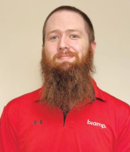 A man with a full reddish-brown beard and short hair, wearing a red collared shirt with biamp. and an Under Armour logo, smiles softly against a plain light background.