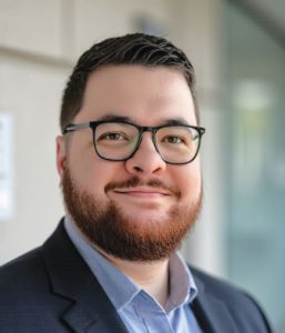 A man with short dark hair, a beard, and glasses is smiling. He is wearing a dark suit jacket over a light blue shirt, standing in front of a blurred background indoors.