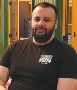 A man with short dark hair and a beard is sitting indoors, wearing a black Nicks Pizza t-shirt. The background features green and yellow paneling and glass doors.