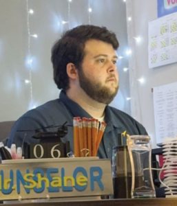 A man with short dark hair and a beard sits at a desk with pencils, a calendar, and office supplies. Counselor and the name Shane are visible on a desk sign. White string lights hang in the background.