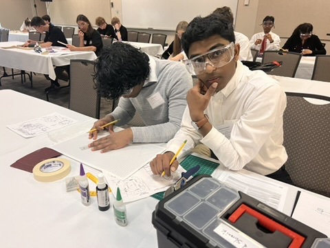 Two students sit at a table working on a project with papers, drawing tools, and craft supplies. One student looks at the camera wearing safety goggles, while the other is focused on drawing. Other students are visible in the background.