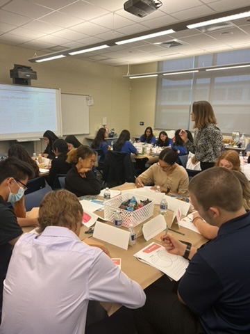 A classroom with students sitting in groups around tables, engaging in a writing or drawing activity. An instructor stands nearby, interacting with students. Papers, name cards, and supplies are spread out on the tables.