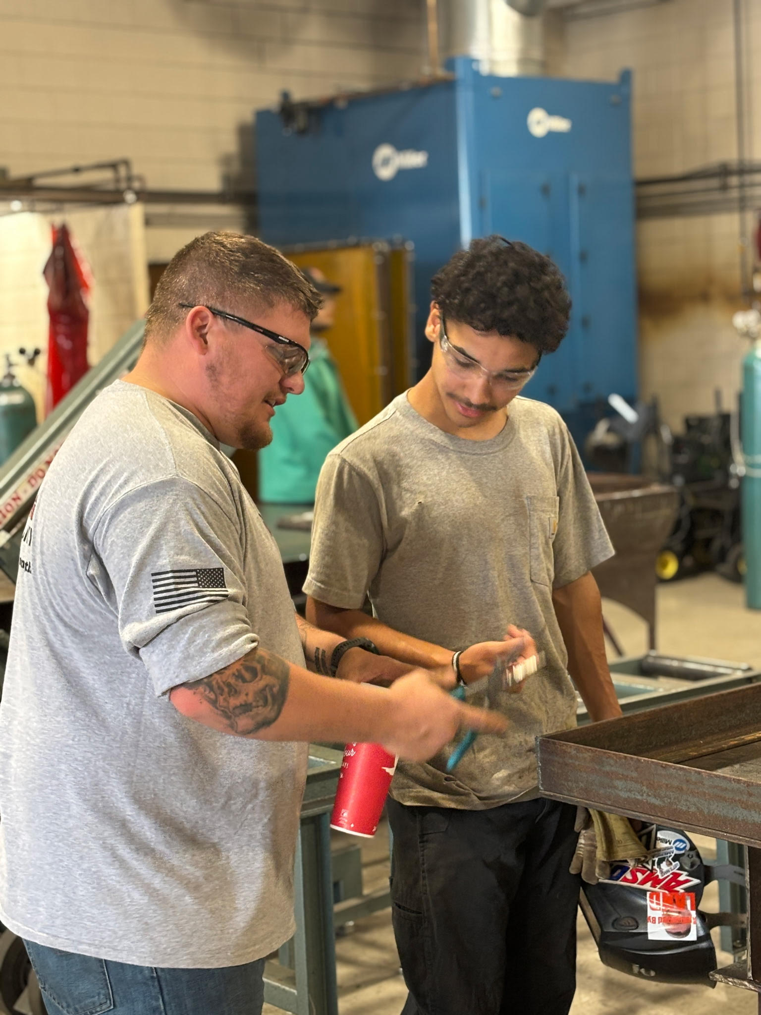Two men in safety glasses work together in a workshop, discussing and examining metal objects. One is holding a tool while the other listens attentively. Workshop equipment and tanks are visible in the background.