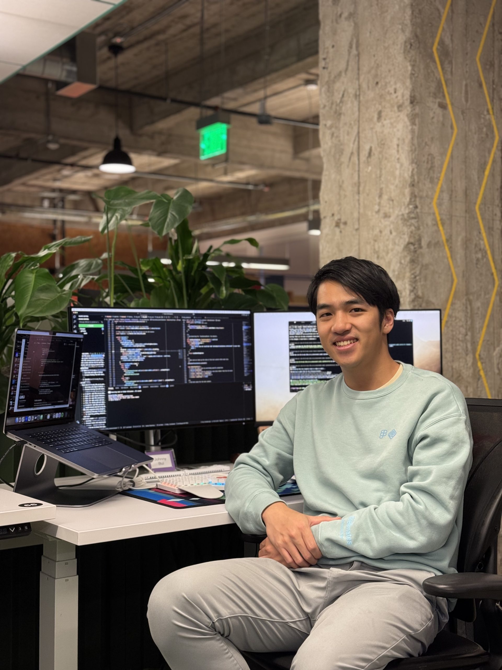 A person sits at a desk with three monitors displaying code in a modern office. They are smiling, wearing a light blue sweatshirt, and surrounded by plants and industrial-style decor.
