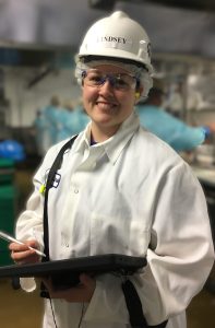 A smiling worker in safety glasses, hair net, white coat, and hard hat labeled Lindsey holds a tablet and stylus in a food processing facility.