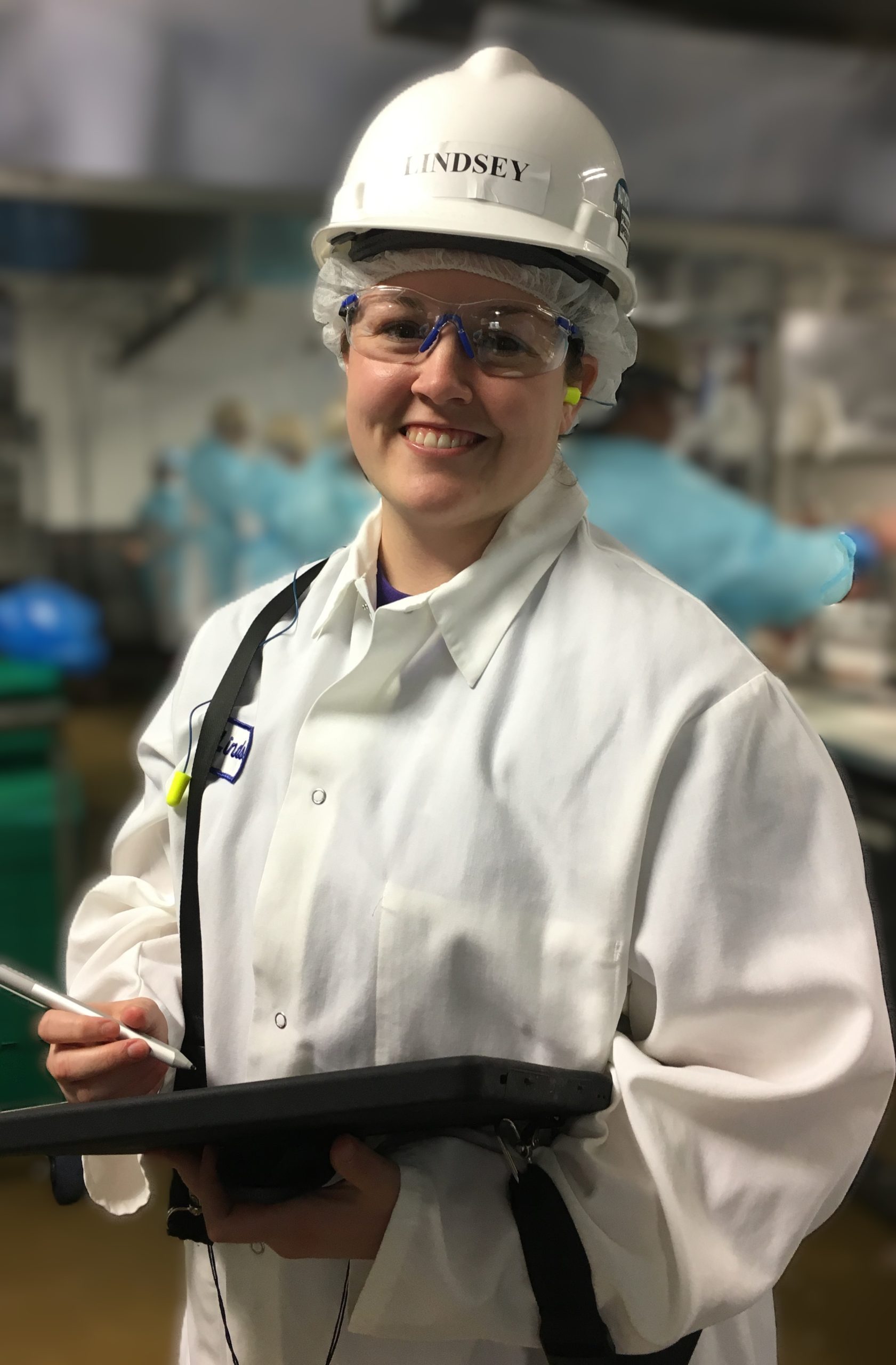 A smiling worker in safety glasses, hair net, white coat, and hard hat labeled Lindsey holds a tablet and stylus in a food processing facility.